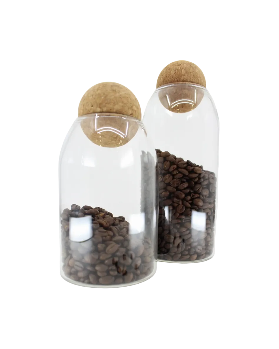 A clear glass coffee canister with a cork lid, filled with coffee beans. Another canister is shown in the background, and there are a few coffee beans scattered in front of the foreground canister.
